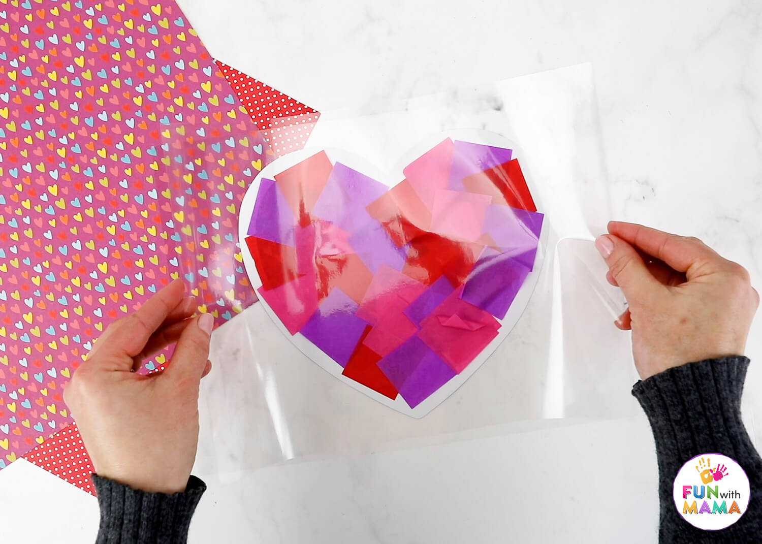 A self-laminating sheet being placed on top of a heart frame filled with pink, red and purple tissue paper squares